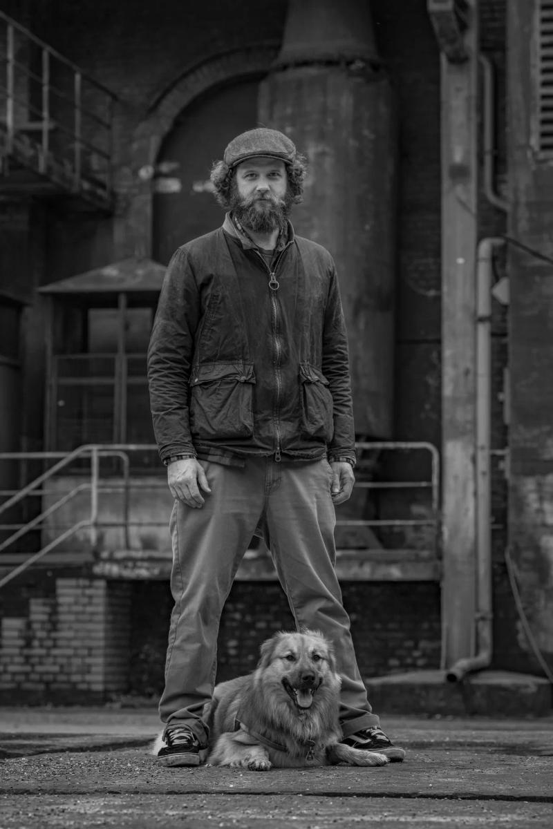 Ein Mann steht mit seinem liegenden Hund vor einem alten Gebäude, in Schwarz-Weiß.
