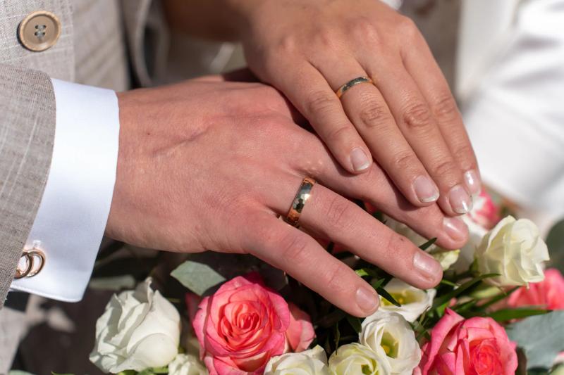Die Hände eines Hochzeitspaars mit Eheringen liegen auf einem Blumenstrauß.