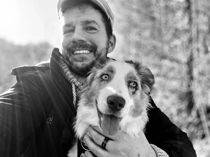 Markus lächelt und hält seine Australian-Shepherd-Hündin Floki im Arm, fotografiert in Schwarz-Weiß.