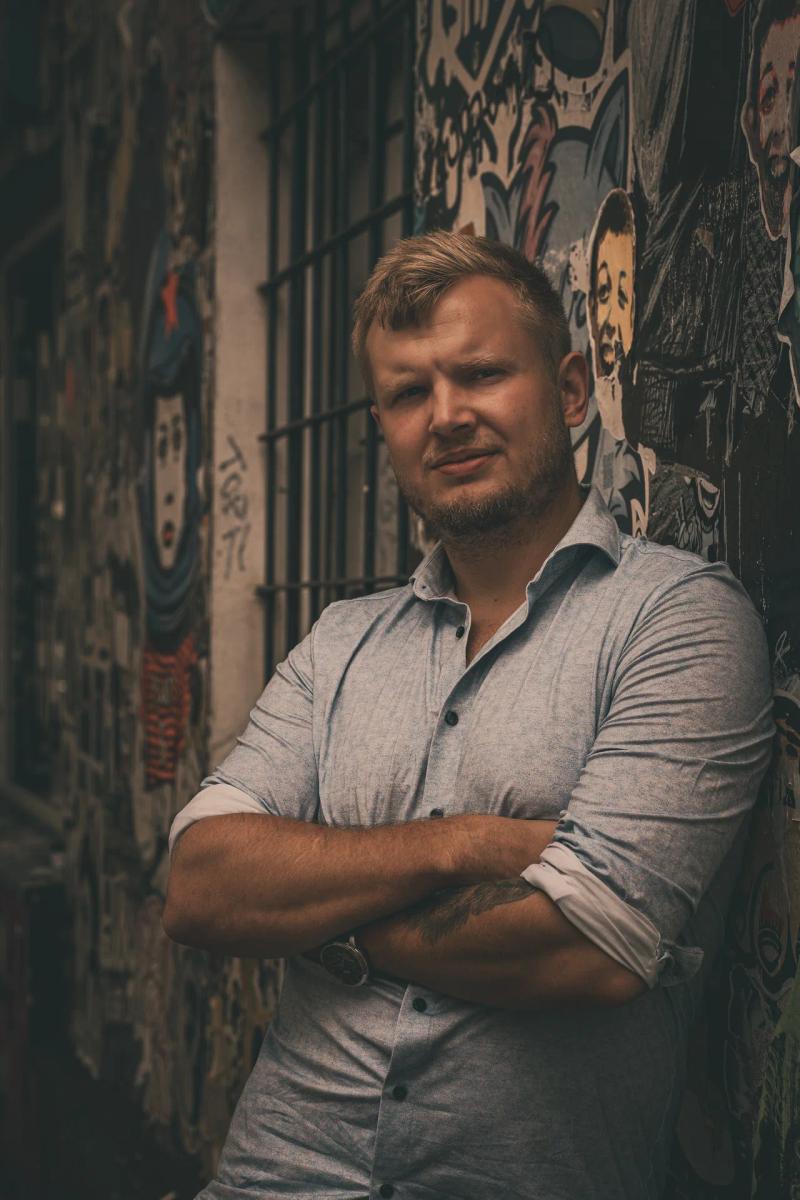 Ein Mann mit verschränkten Armen lehnt an einer mit Graffiti bedeckten Wand.