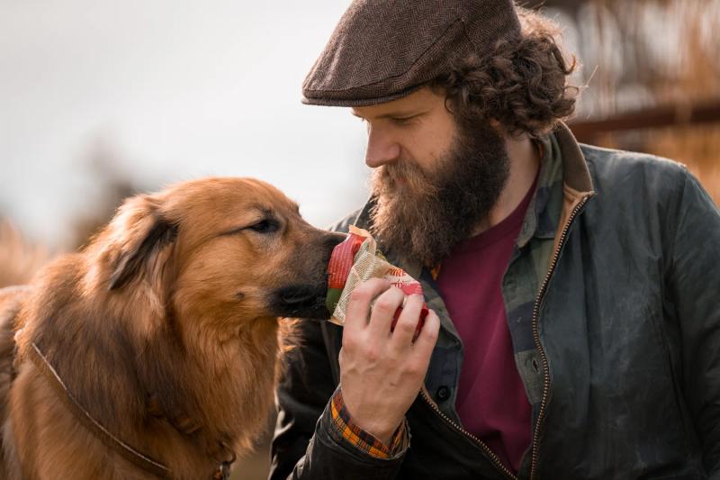 Ein bärtiger Mann mit Mütze hält seinen Golden Retriever nah an sein Gesicht.