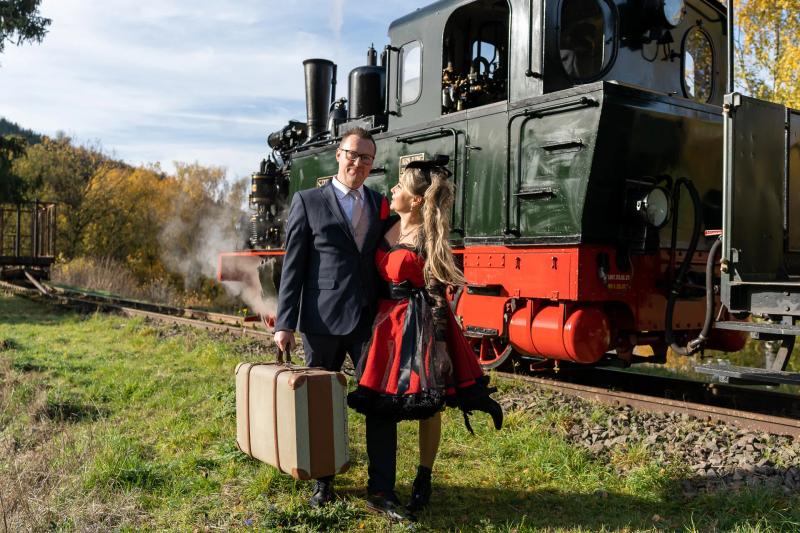 Ein Hochzeitspaar steht mit Koffern vor einer historischen Dampflokomotive.