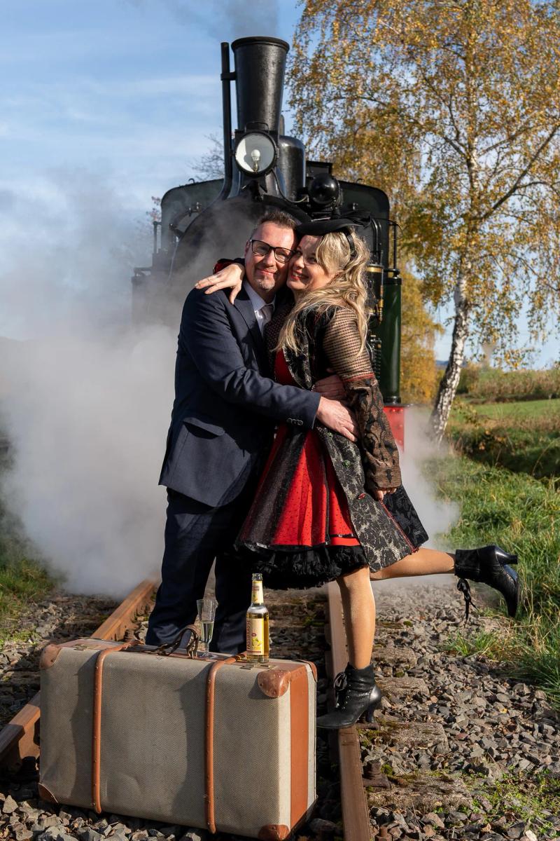 Ein Hochzeitspaar umarmt sich neben einer dampfenden Lokomotive.