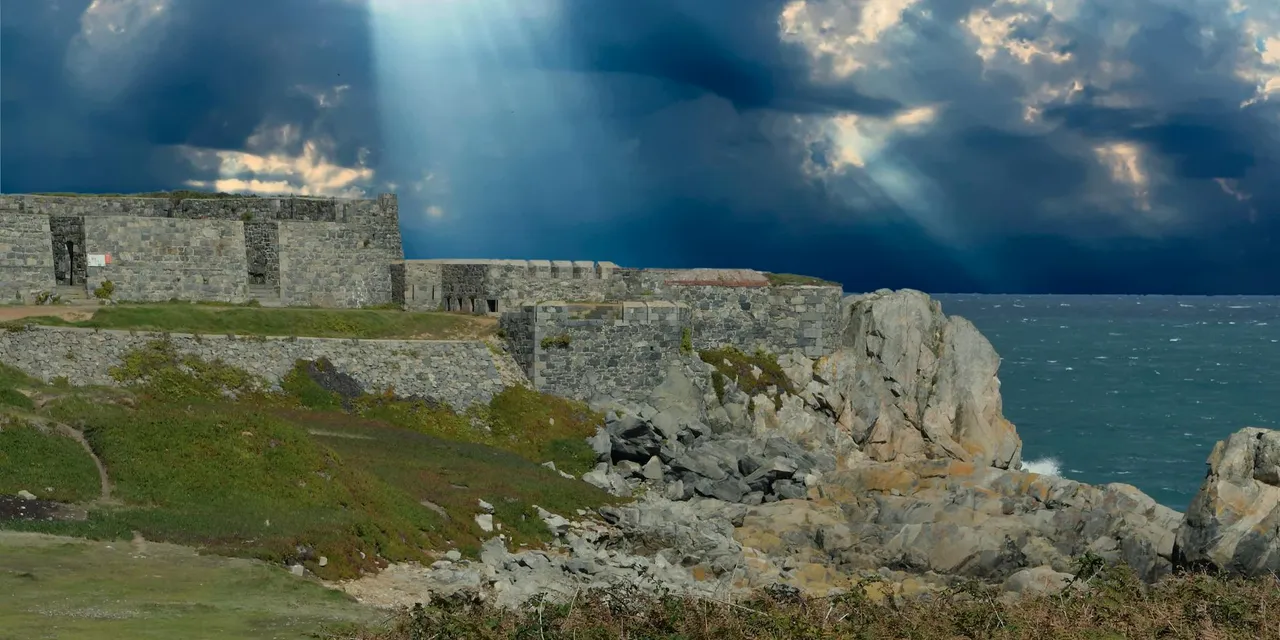 Rugged fort along Guernsey's rocky coast with dramatic clouds and sunrays breaking through.
