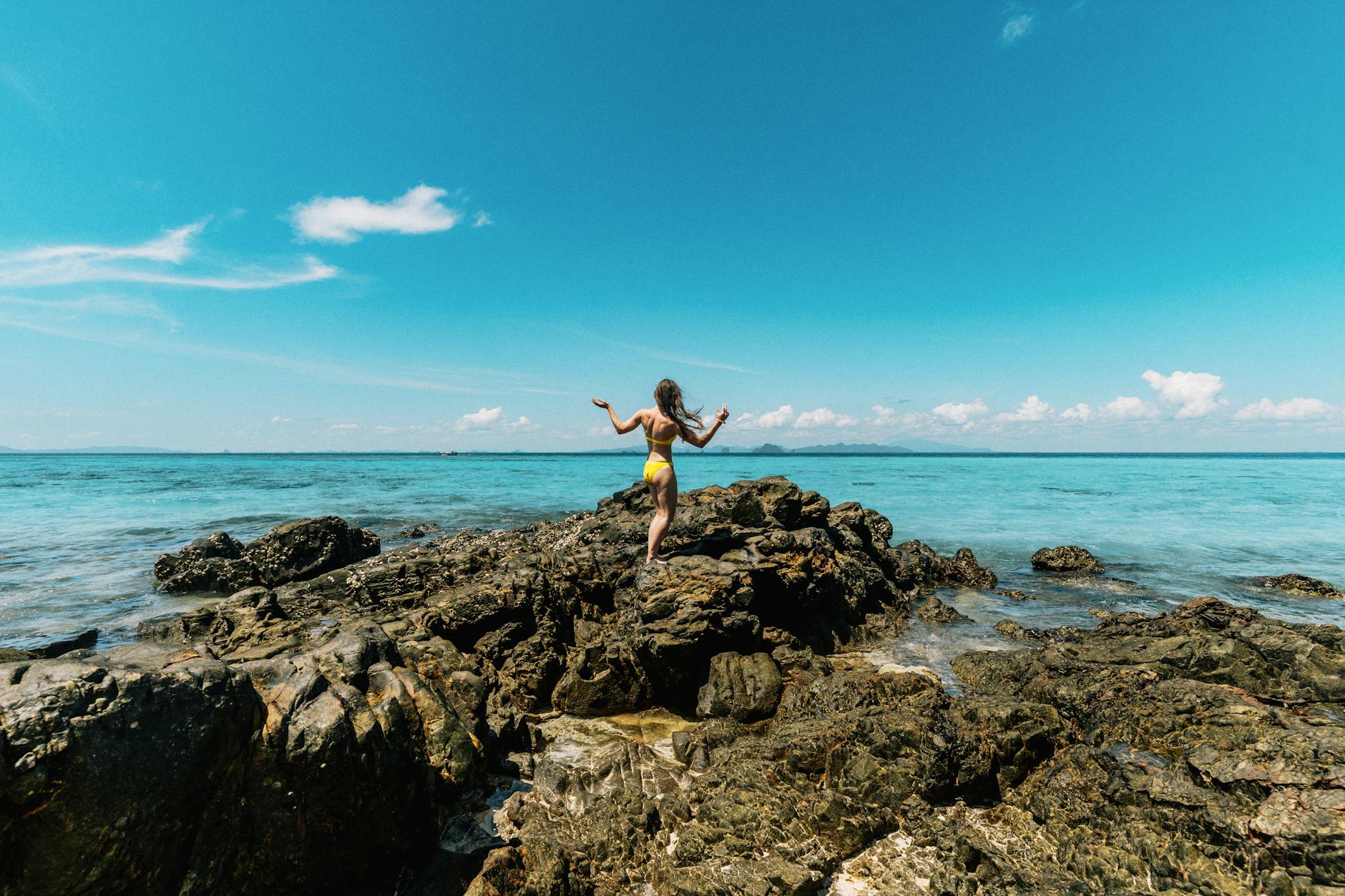 A woman in a yellow swimsuit stands on rocky coast in Ao Nang, Krabi, Thailand under a clear blue sky.
