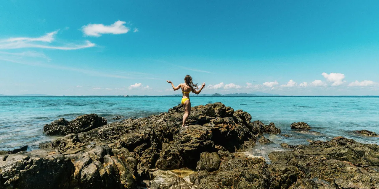 A woman in a yellow swimsuit stands on rocky coast in Ao Nang, Krabi, Thailand under a clear blue sky.