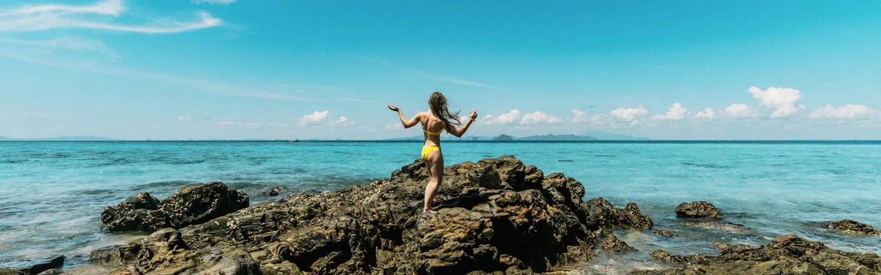 A woman in a yellow swimsuit stands on rocky coast in Ao Nang, Krabi, Thailand under a clear blue sky.