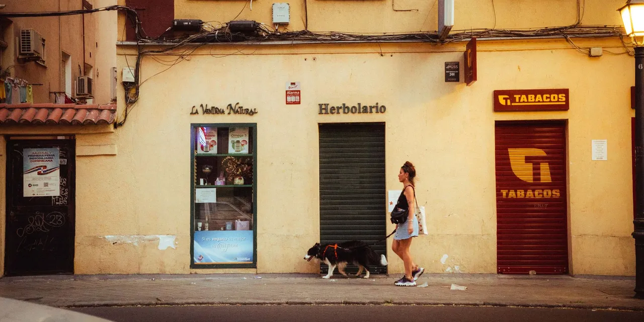 A woman walking her dog past storefronts on a street in Madrid.