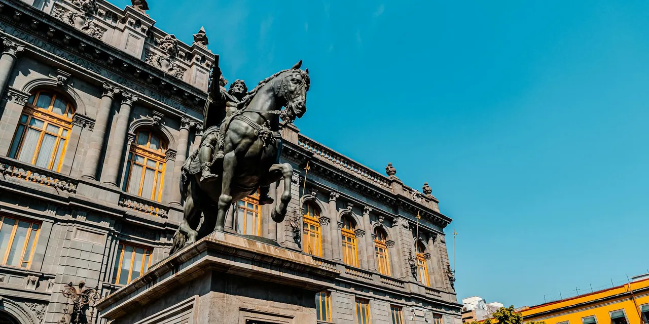 Low-angle shot of a historic building and statue in Mexico City with a clear blue sky.