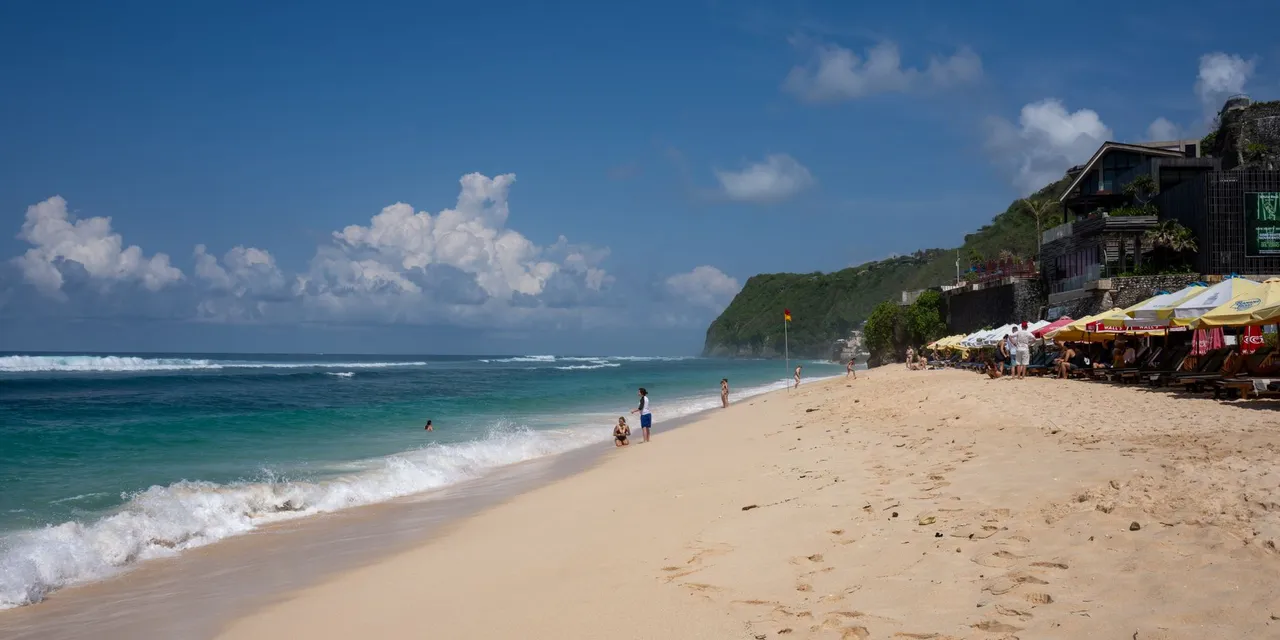 A vibrant beach scene in Bali, Indonesia with clear blue skies, ocean waves, and beachgoers enjoying the sun.