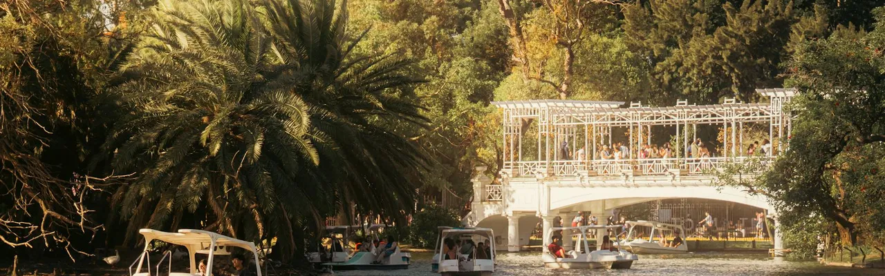 Boaters enjoy a sunny day under a white bridge in a lush park.