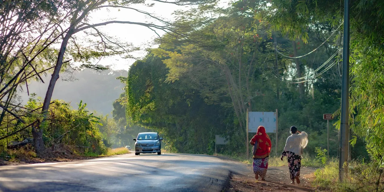 A serene road in Dembéni, Mayotte, with two pedestrians walking along lush greenery.
