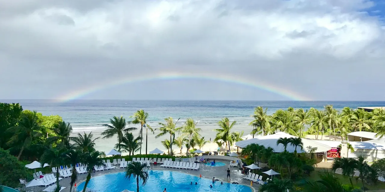 Aerial view of a tropical resort pool in Tumon, Guam with a vibrant rainbow above the ocean, showcasing palm trees and a summer ambiance.