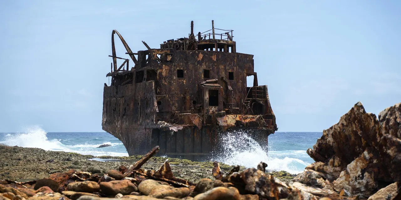 A rusting shipwreck on Curaçao's rocky coast with waves crashing, under a clear blue sky.