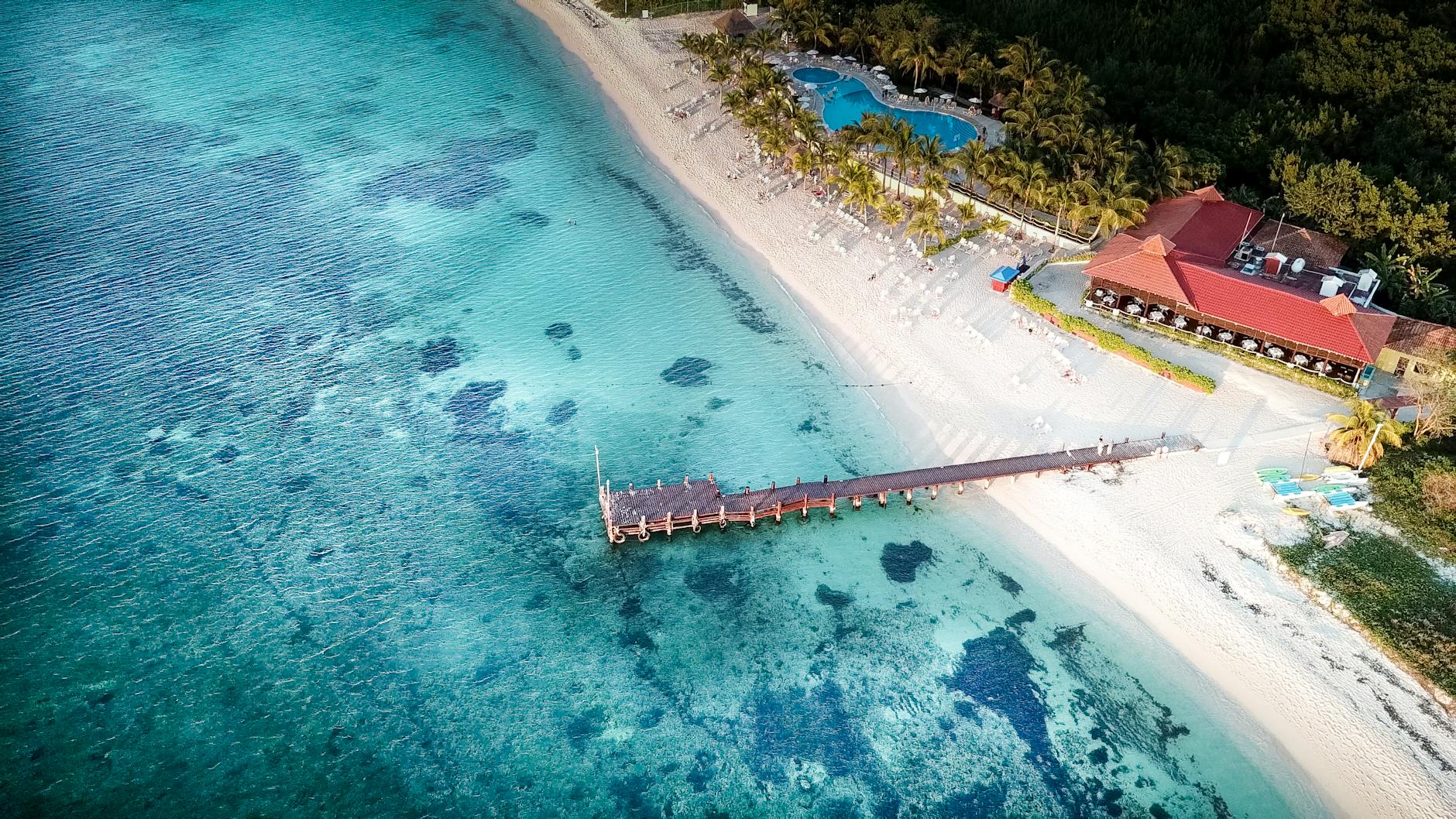 Aerial shot of a tropical beach with a pier, turquoise waters, and a resort in Cozumel.