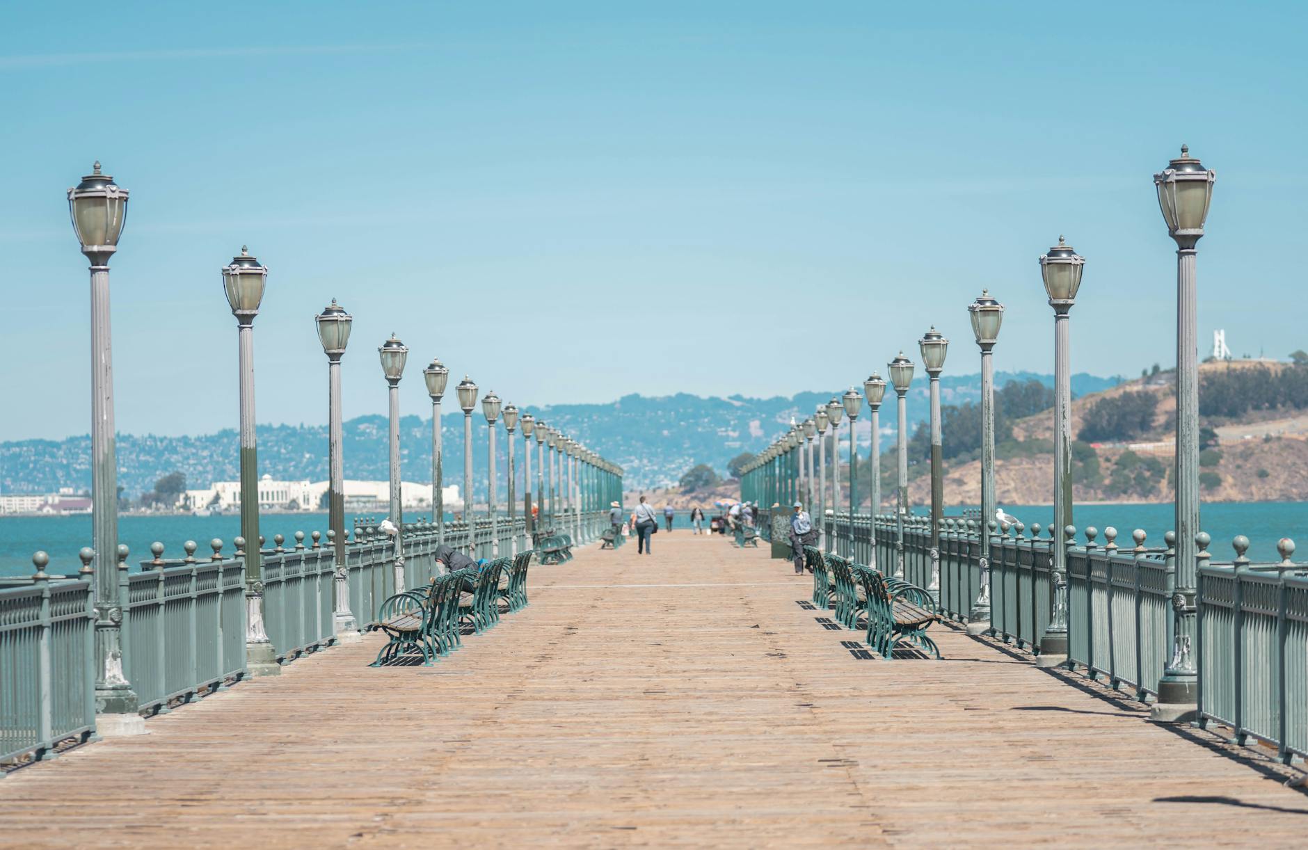 A picturesque wooden pier lined with lampposts, offering views of a serene bay and distant hills.