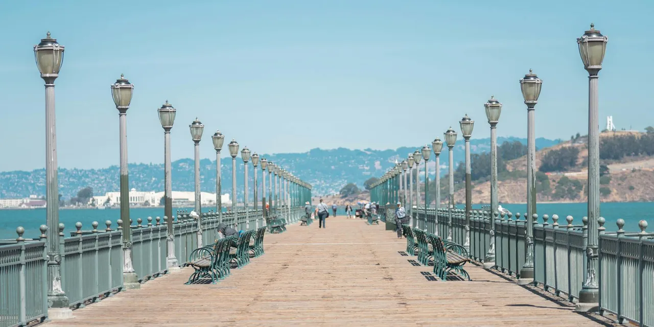 A picturesque wooden pier lined with lampposts, offering views of a serene bay and distant hills.