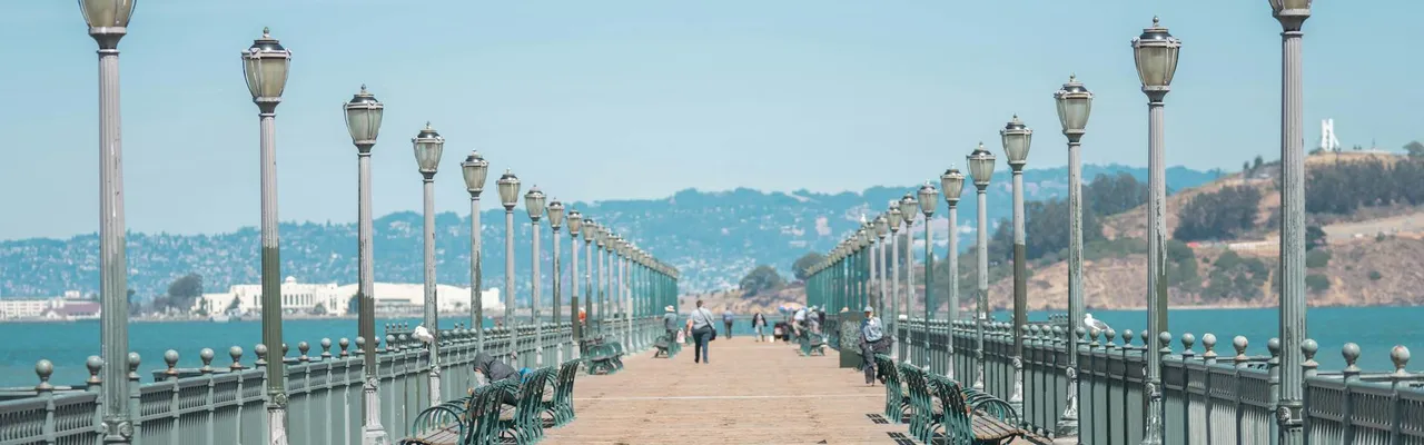 A picturesque wooden pier lined with lampposts, offering views of a serene bay and distant hills.
