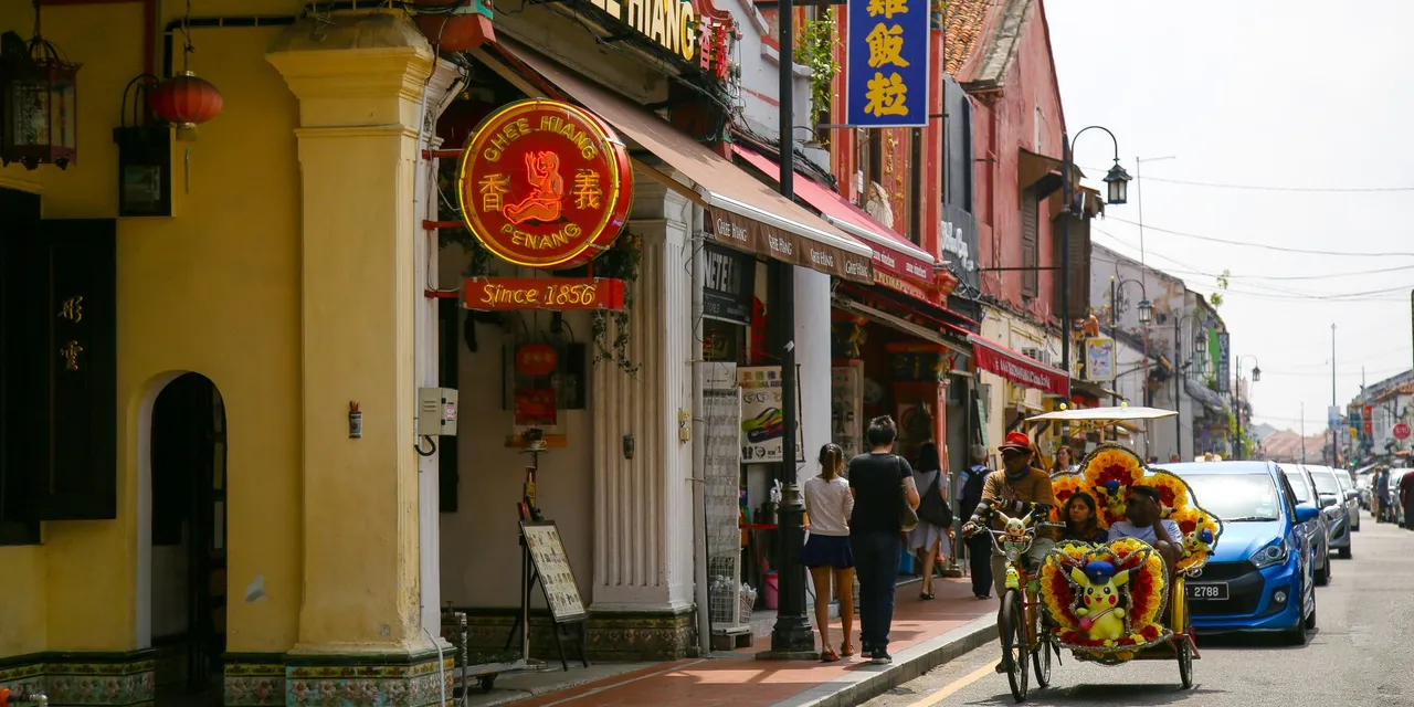 Vibrant trishaw passing traditional shops on a sunny Malacca street.