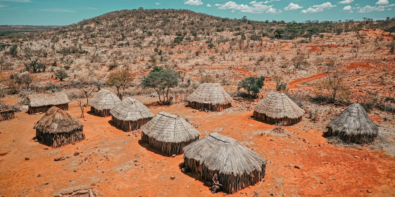 Aerial view of traditional huts in Angola set against a dry, rugged landscape under a bright blue sky.