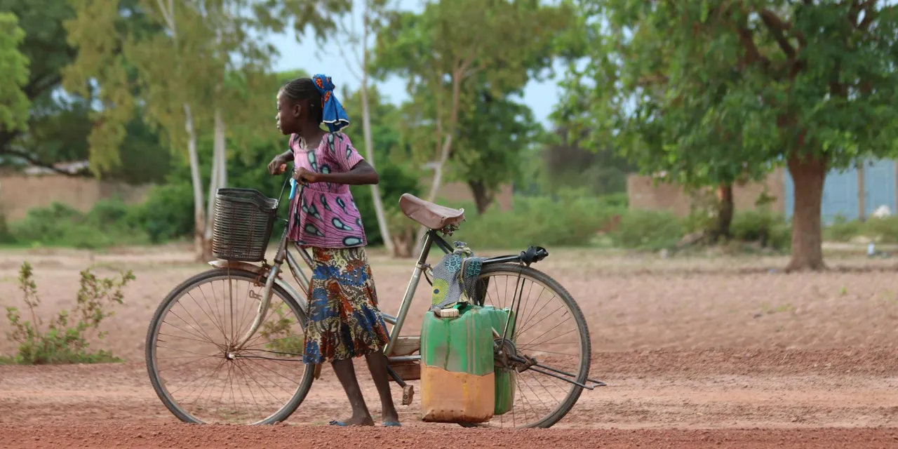 A young girl pushing a bicycle with containers in a rural field in Burkina Faso.