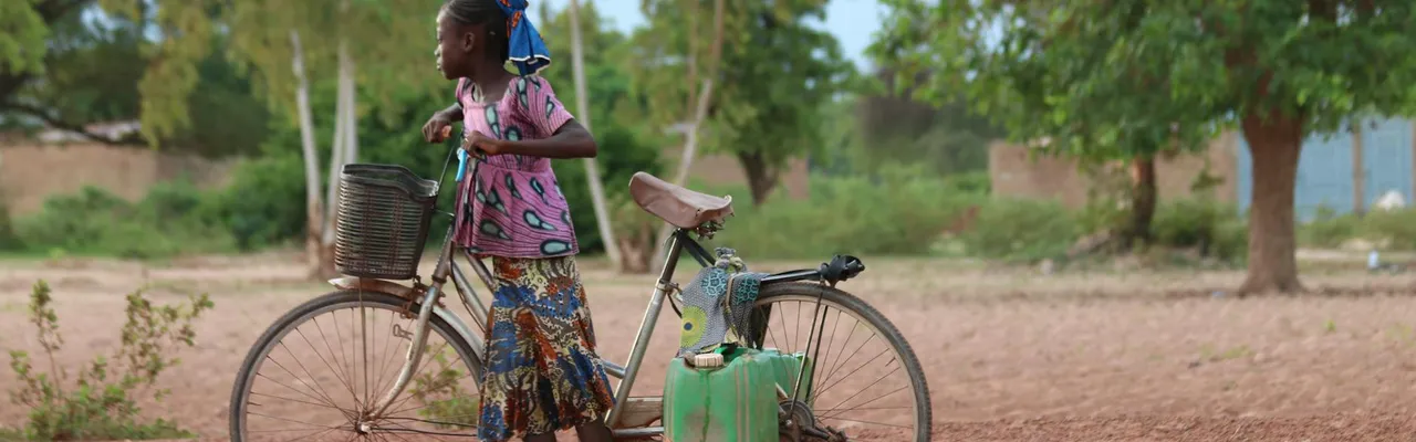 A young girl pushing a bicycle with containers in a rural field in Burkina Faso.