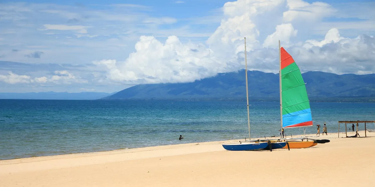 Vibrant sailboats rest on a sunny Lake Malawi beach with mountains in the background.