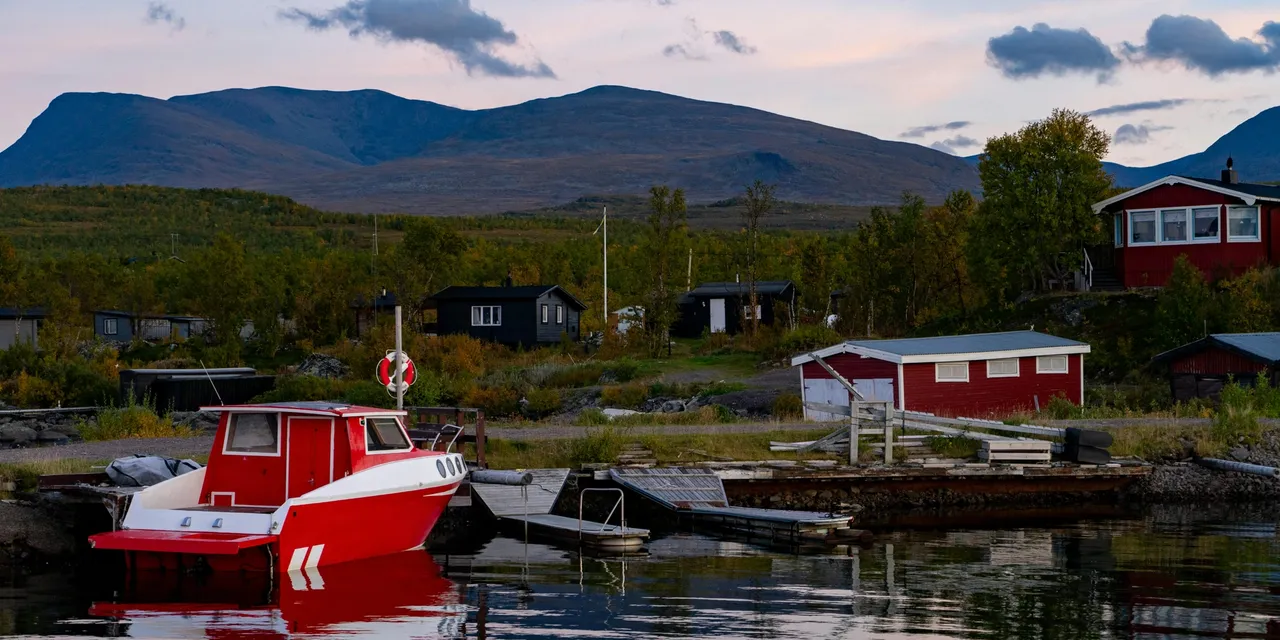 Red boat moored at a quiet Swedish lakeside village with mountains behind, Sweden