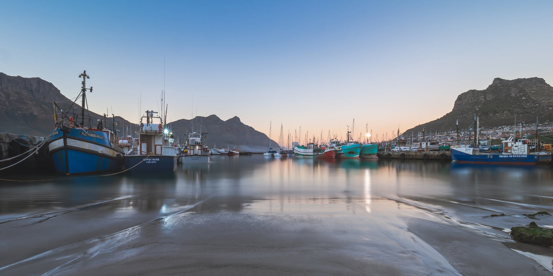 Captured at sunset, this tranquil scene showcases fishing boats reflecting on the calm waters of Hout Bay Harbor.