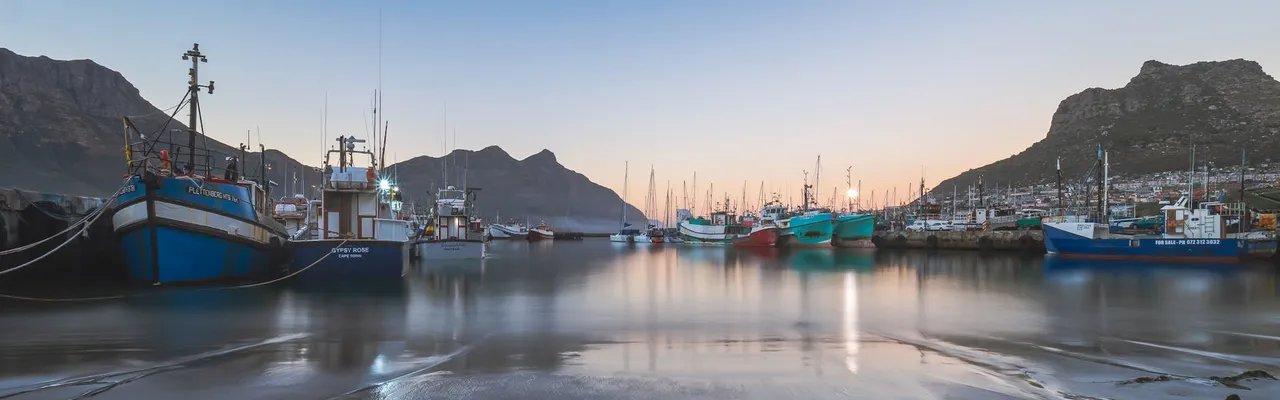 Captured at sunset, this tranquil scene showcases fishing boats reflecting on the calm waters of Hout Bay Harbor.