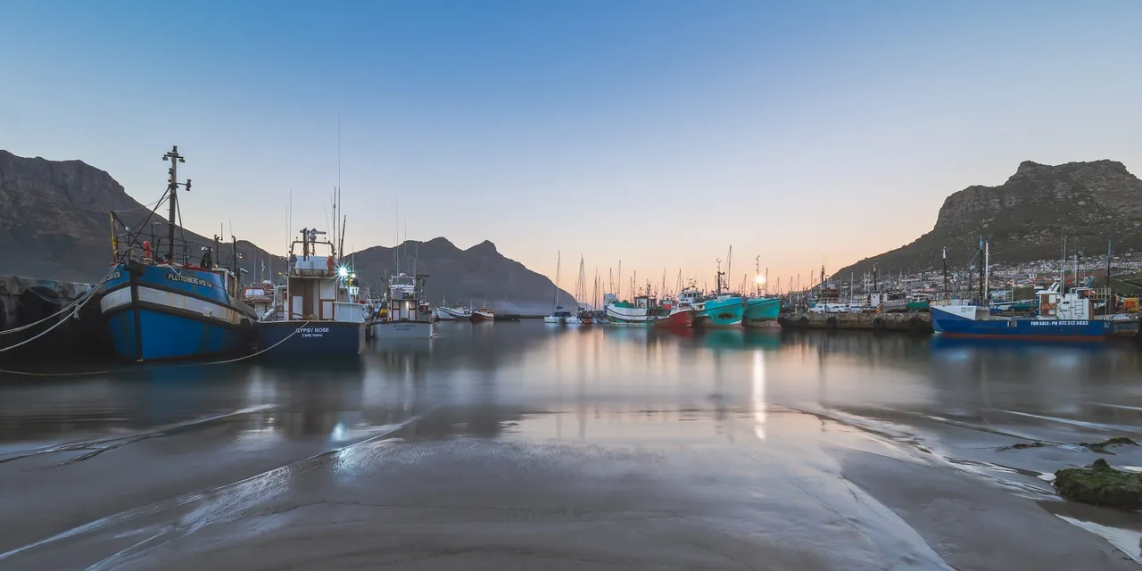 Captured at sunset, this tranquil scene showcases fishing boats reflecting on the calm waters of Hout Bay Harbor.