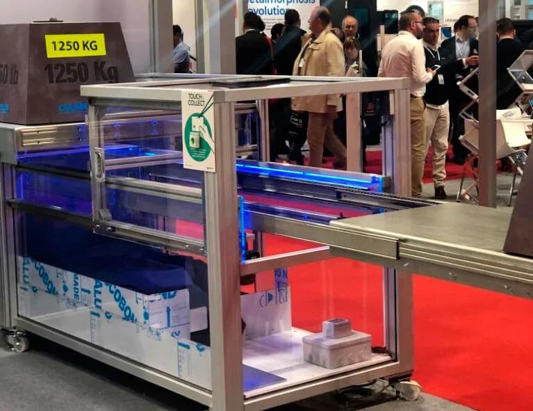 Máquina de embalaje expuesta en un pabellón de feria industrial con visitantes circulando por el pasillo al fondo