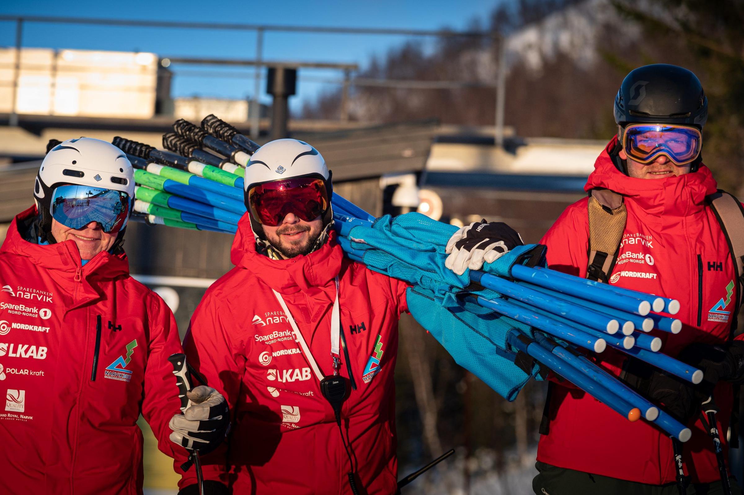 Tre frivillige i røde jakker med hjelm og skibriller bærer slalåmporter i bakken.