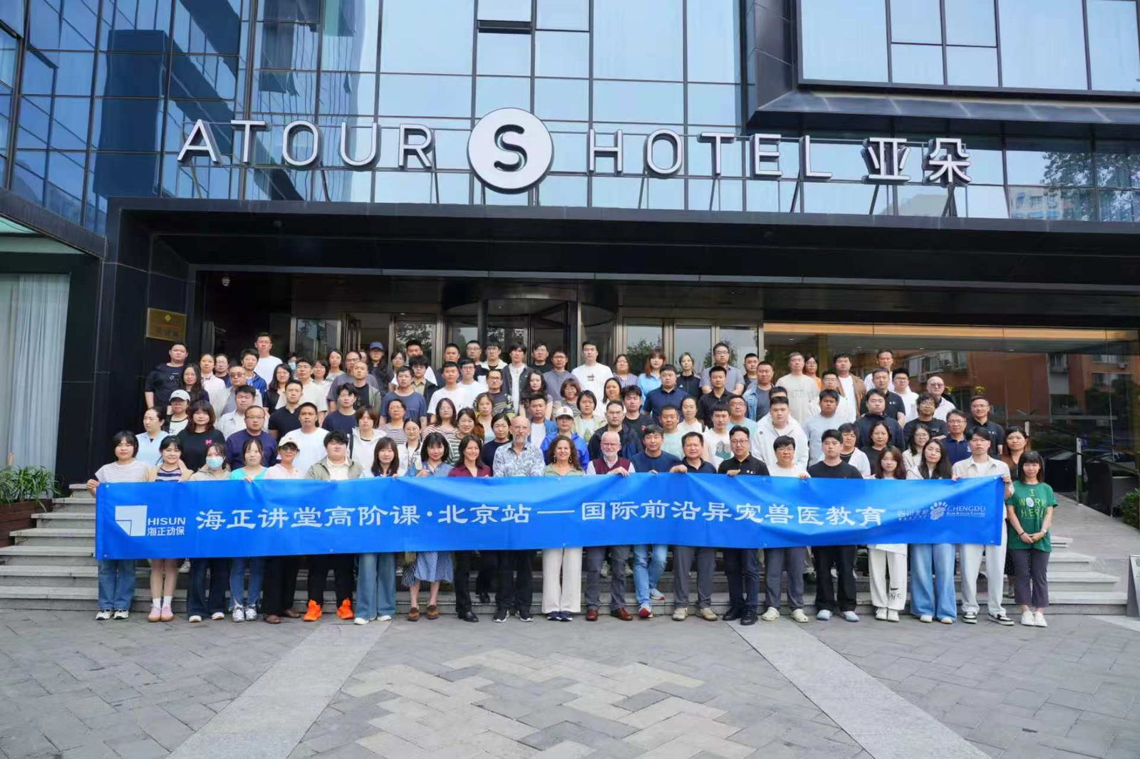A large group of people stand outside the Atour S Hotel, holding a blue banner with Chinese text.