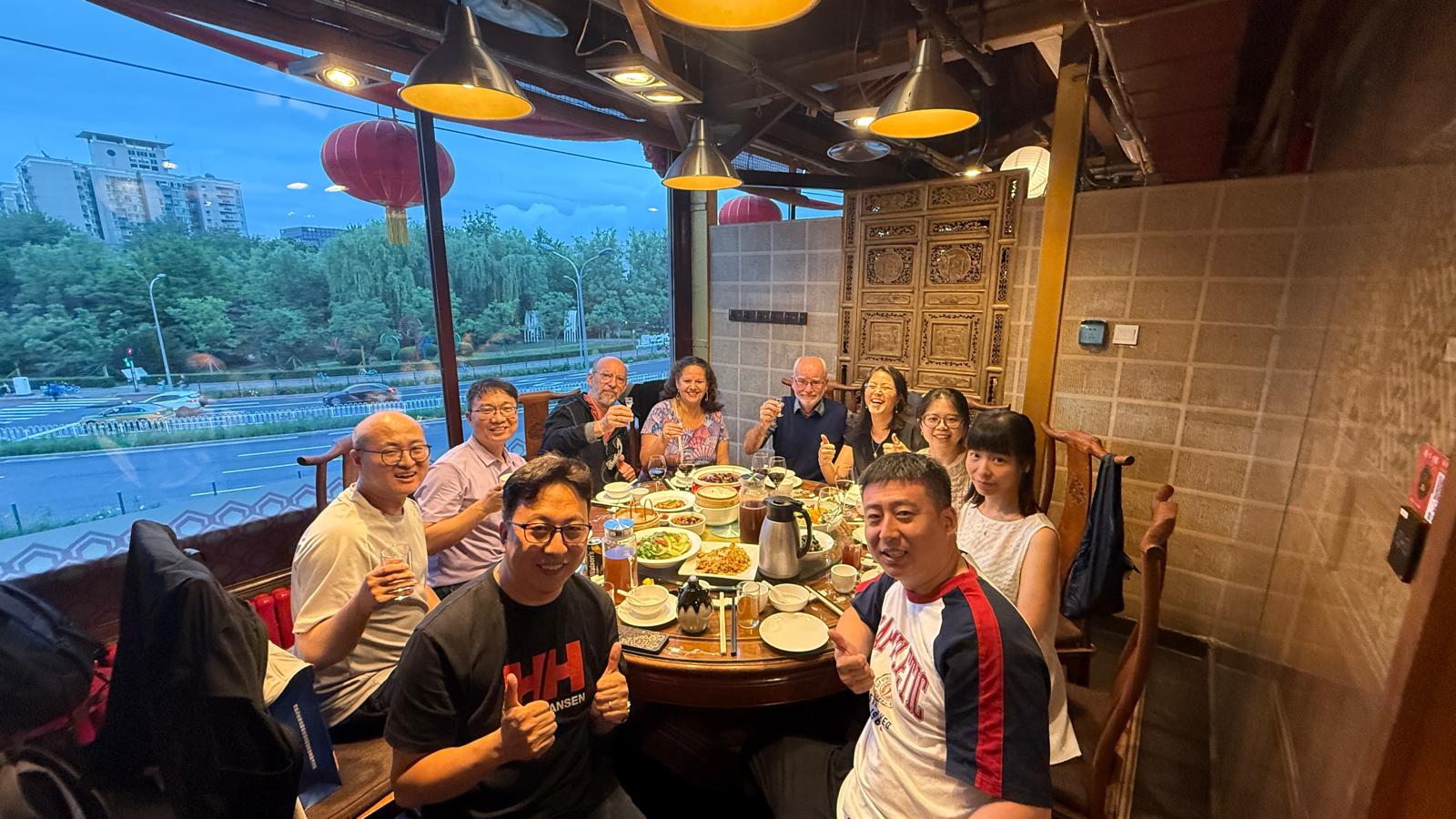 Nine smiling people at a restaurant table, some holding drinks for a toast and others giving thumbs-up.