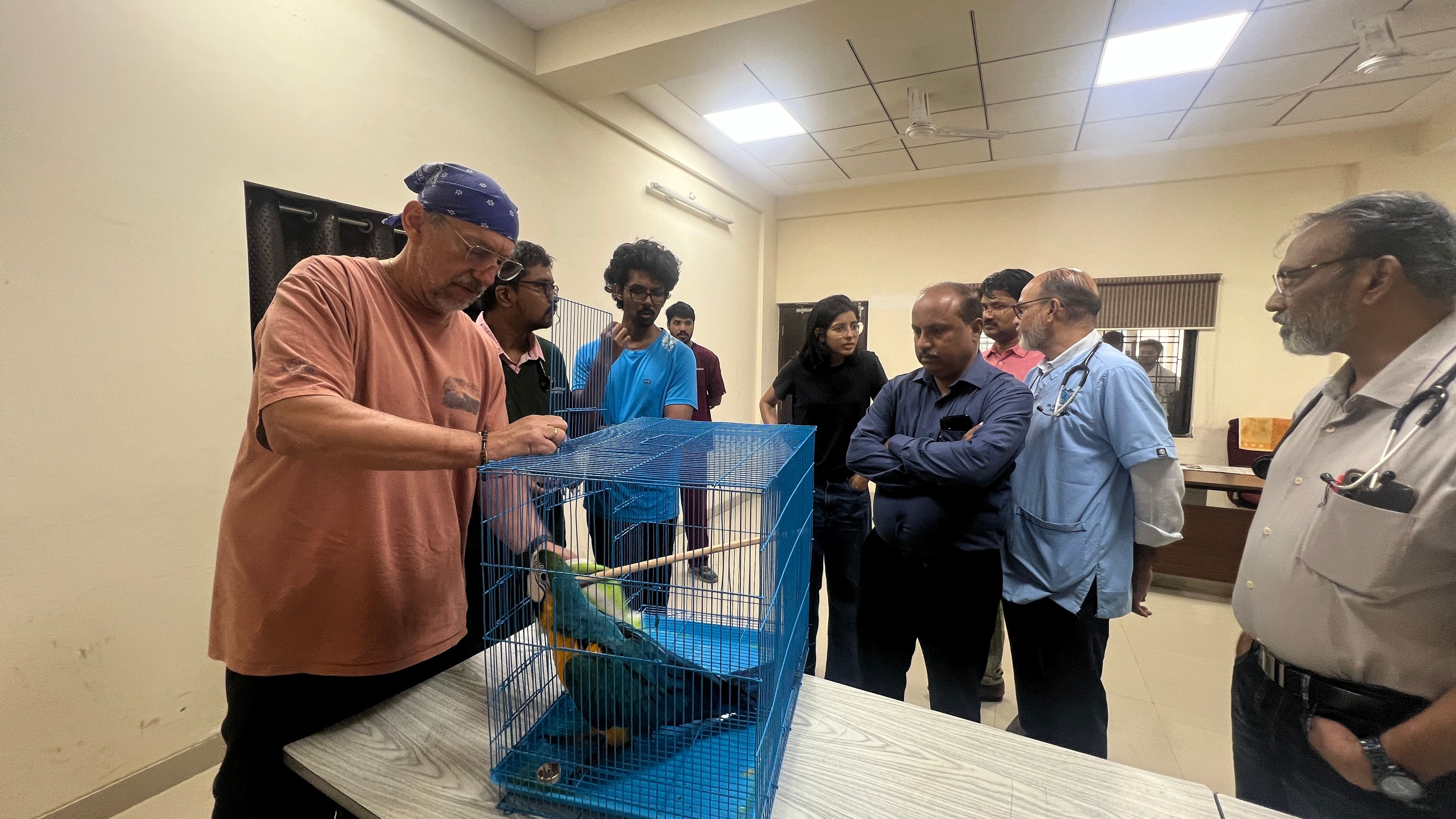 A man interacts with a blue and yellow macaw in a cage, observed by a group of people including veterinary professionals.
