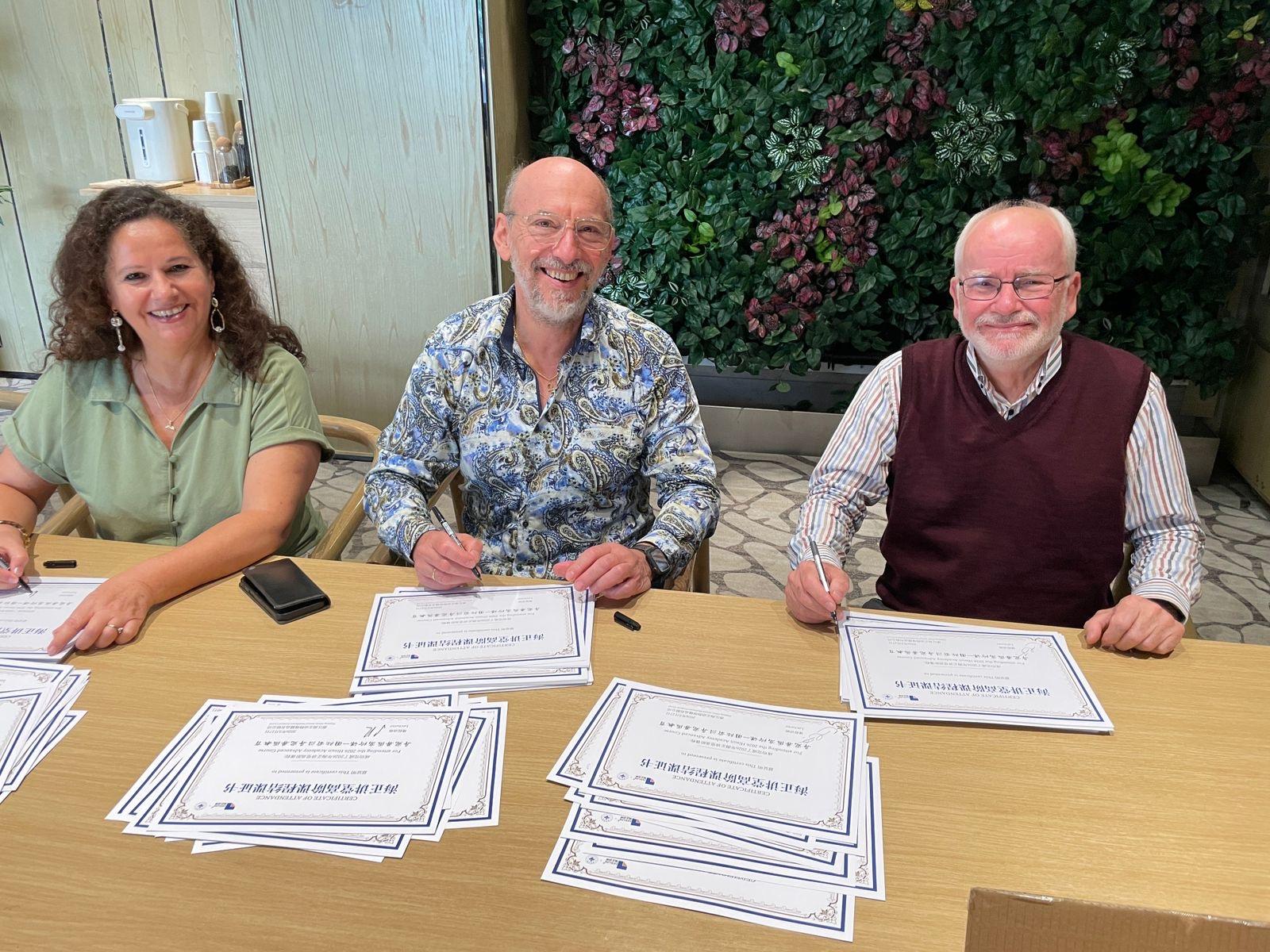 A woman and two men smile at a table with pens and stacks of certificates.