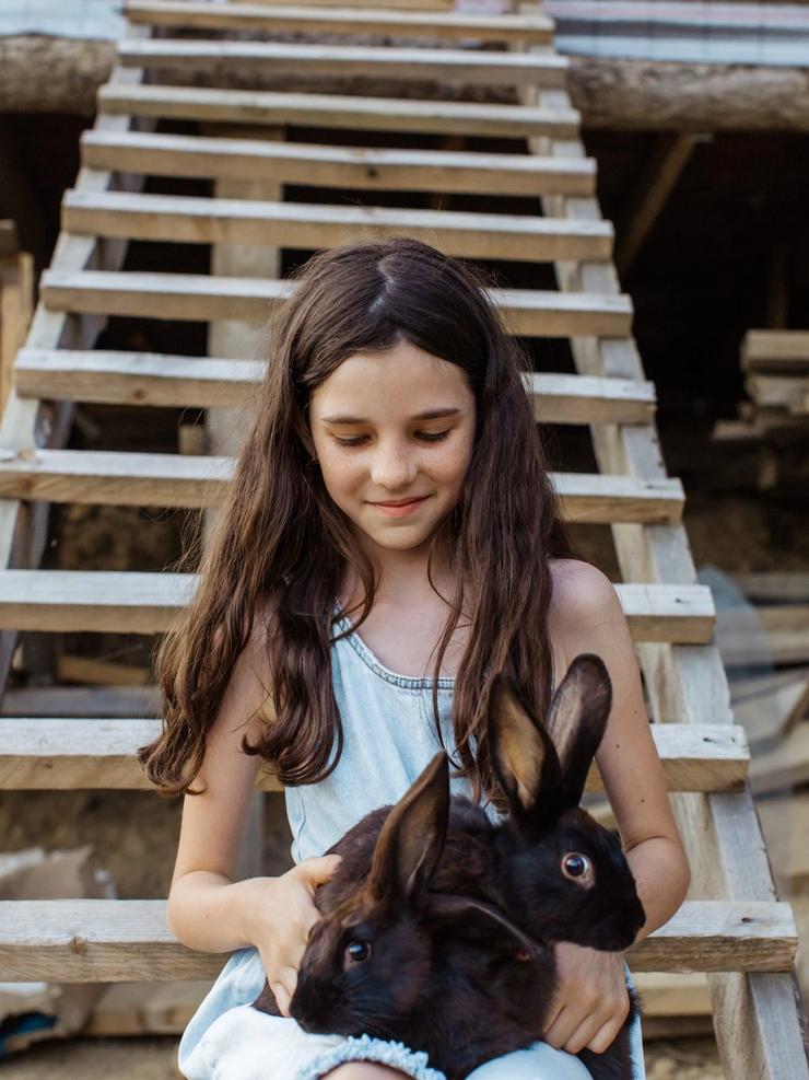 A smiling girl with long hair sits on wooden steps, holding two dark rabbits on her lap.