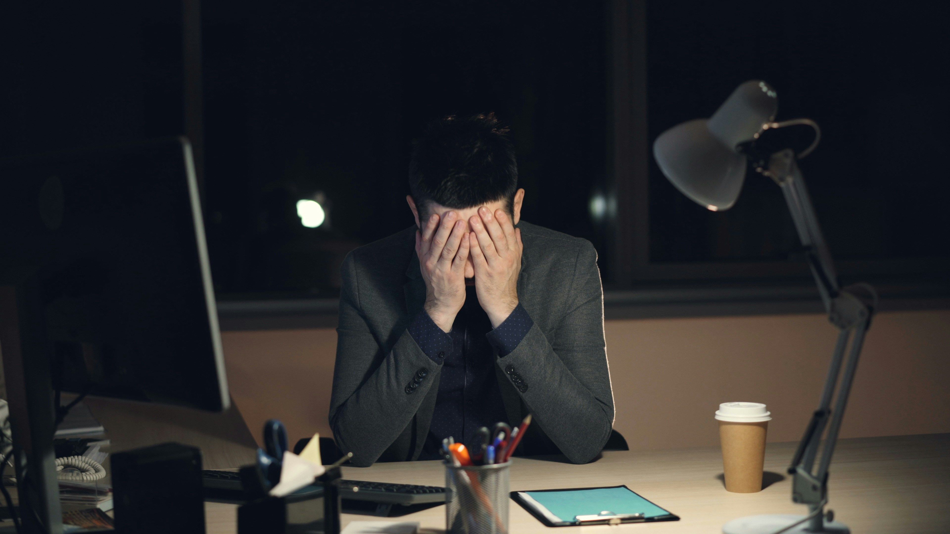 A distressed businessman sits alone at his desk late at night with his face buried in his hands, surrounded by office supplies and a coffee cup, depicting workplace stress and mental health struggles.