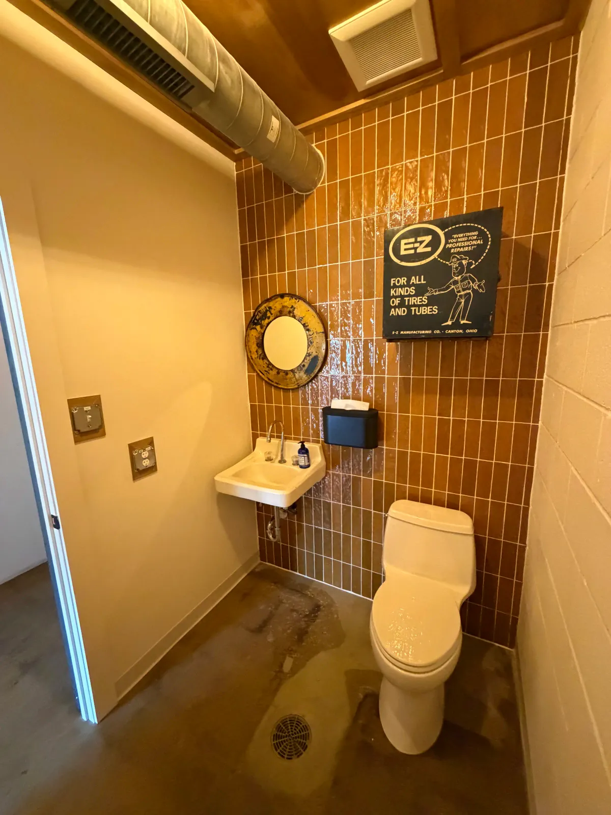 Customer restroom with a vintage EZ For All Kinds of Tires and Tubes sign, brown tile, and a silver vent hood inside Moon Wolf Provisions in Richardson Texas