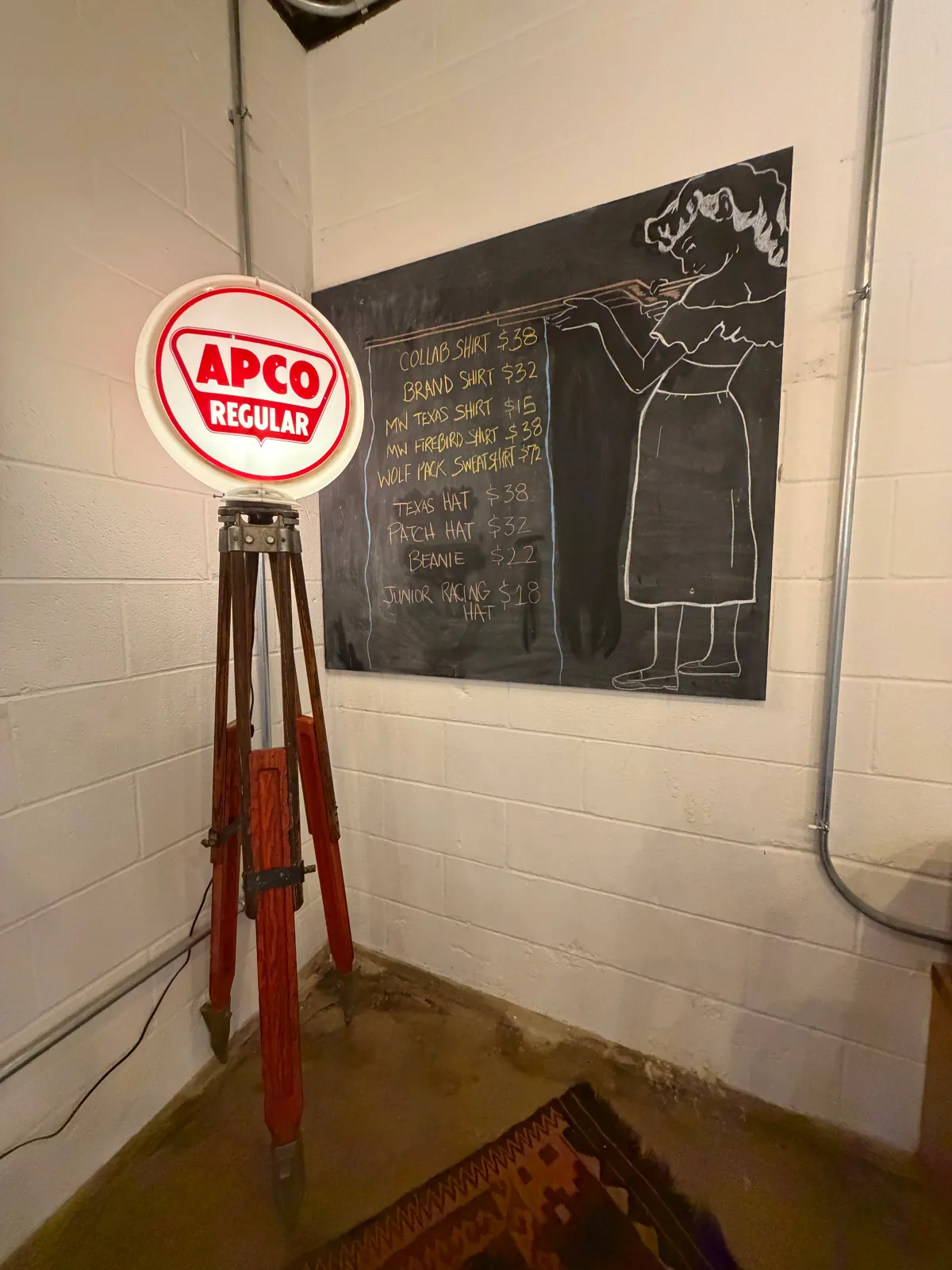 Hand-lettered chalkboard merchandise menu with a vintage APCO Regular gas-pump globe on a wooden tripod inside Moon Wolf Provisions general store in Richardson Texas