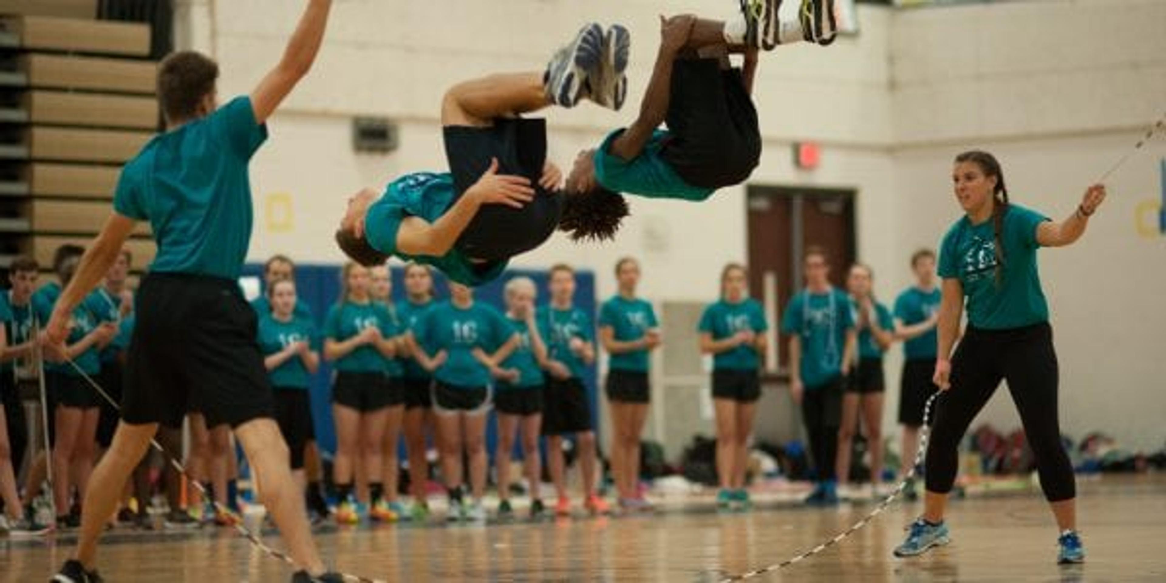 two young adults doing flips as others cheer in the background.