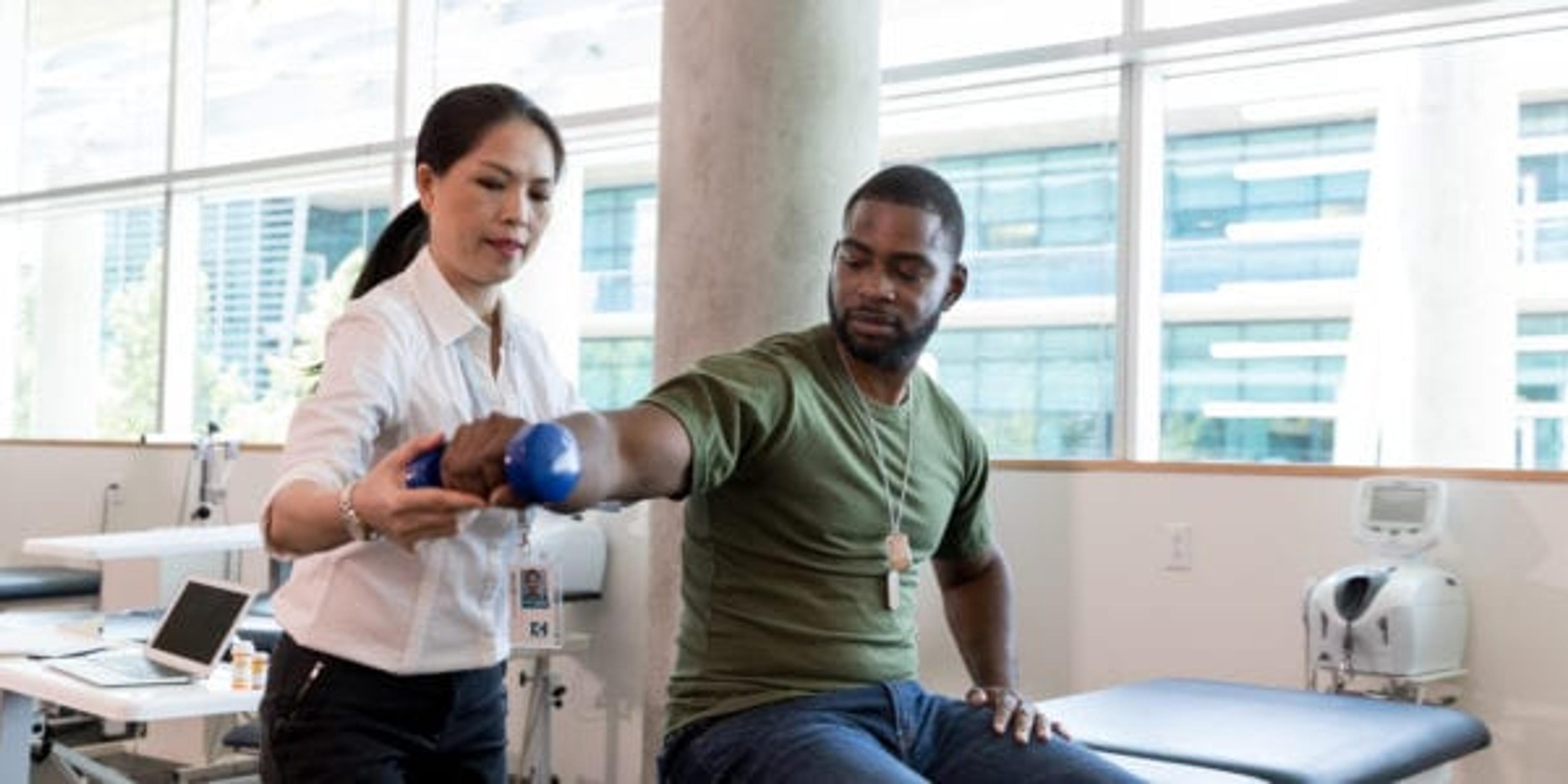 Serious army soldier with injury uses a hand weight during a physical therapy session. A female physical therapist is helping him with an exercise.