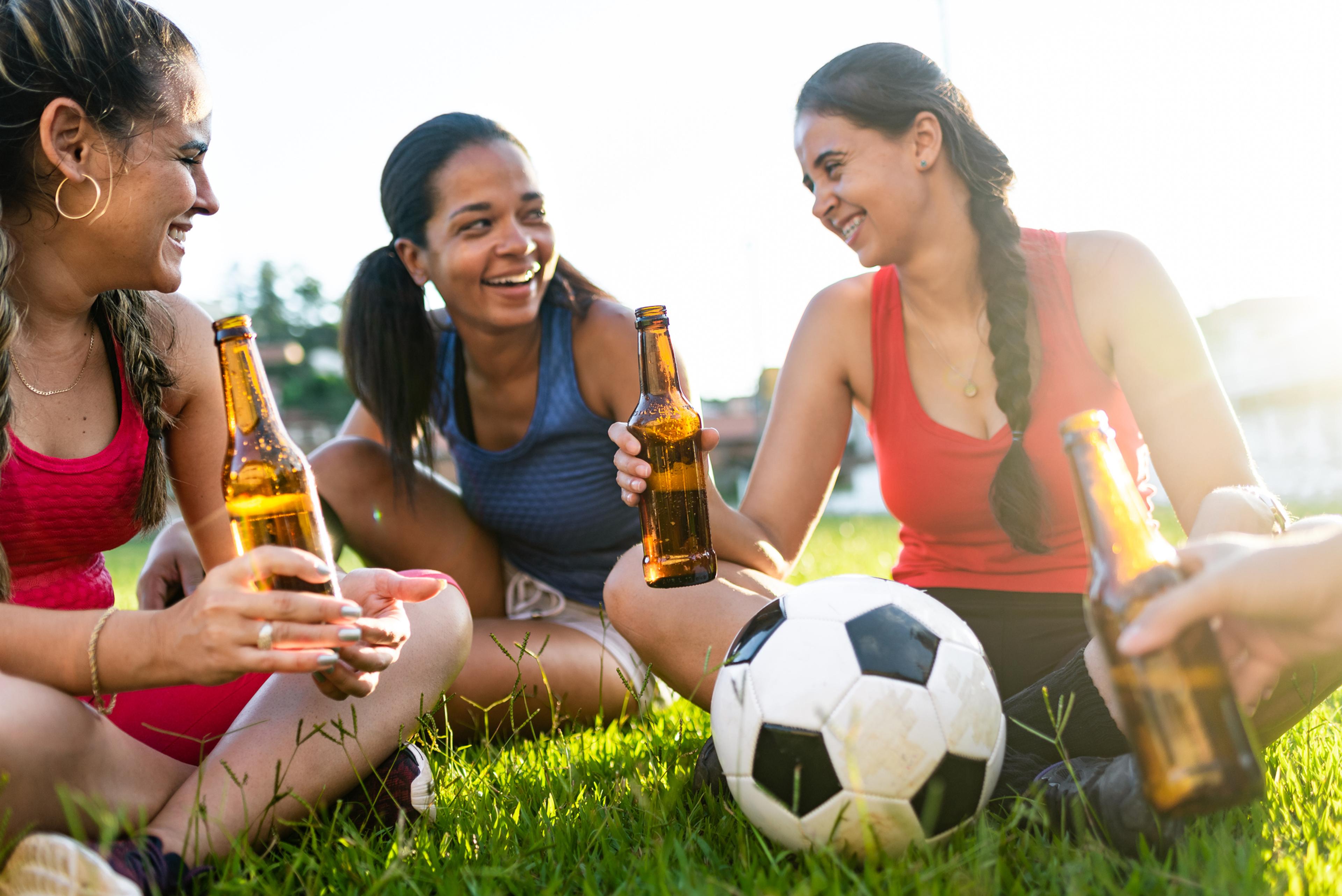 A group of women sitting on a grass field after playing soccer, sharing bottled beers.
