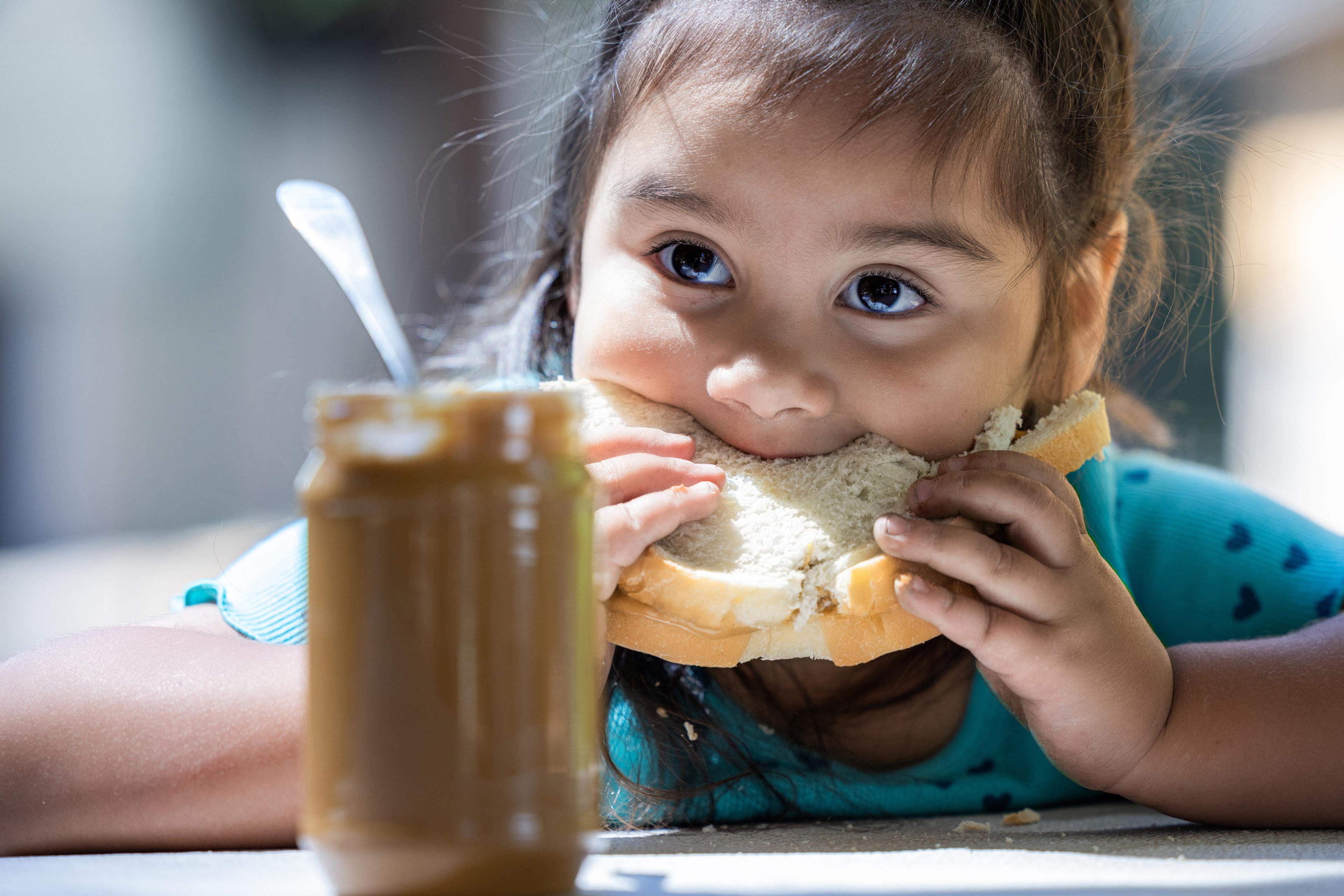 A young girl is eating a sandwich with a jar of peanut butter next to her. The scene is casual and relaxed, with the girl enjoying her snack.