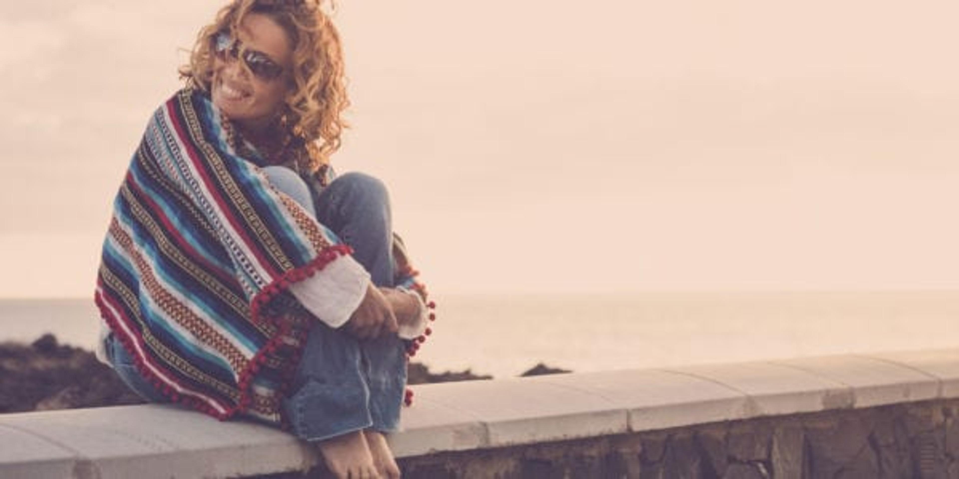 smiling woman cuddled up in a blanket sitting on a bench/ledge overlooking water