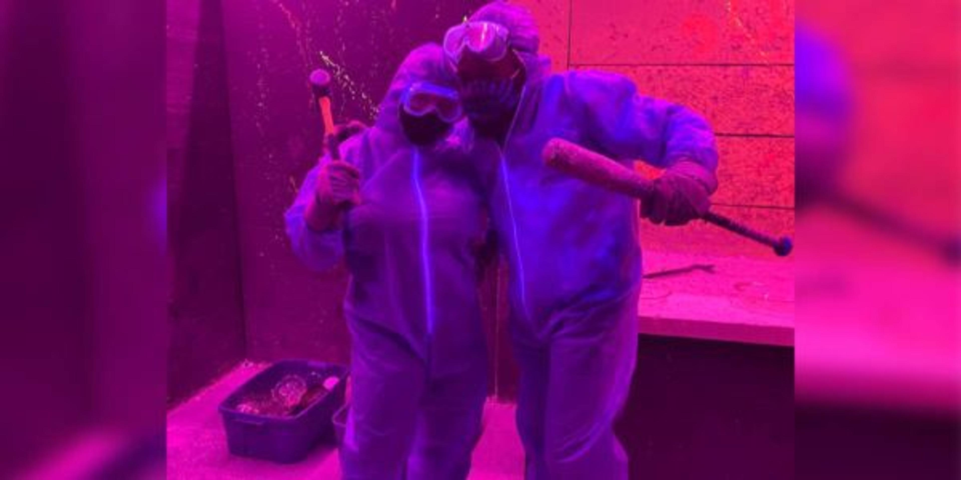 Two people in protective gear pose for a photo inside of a rage room.