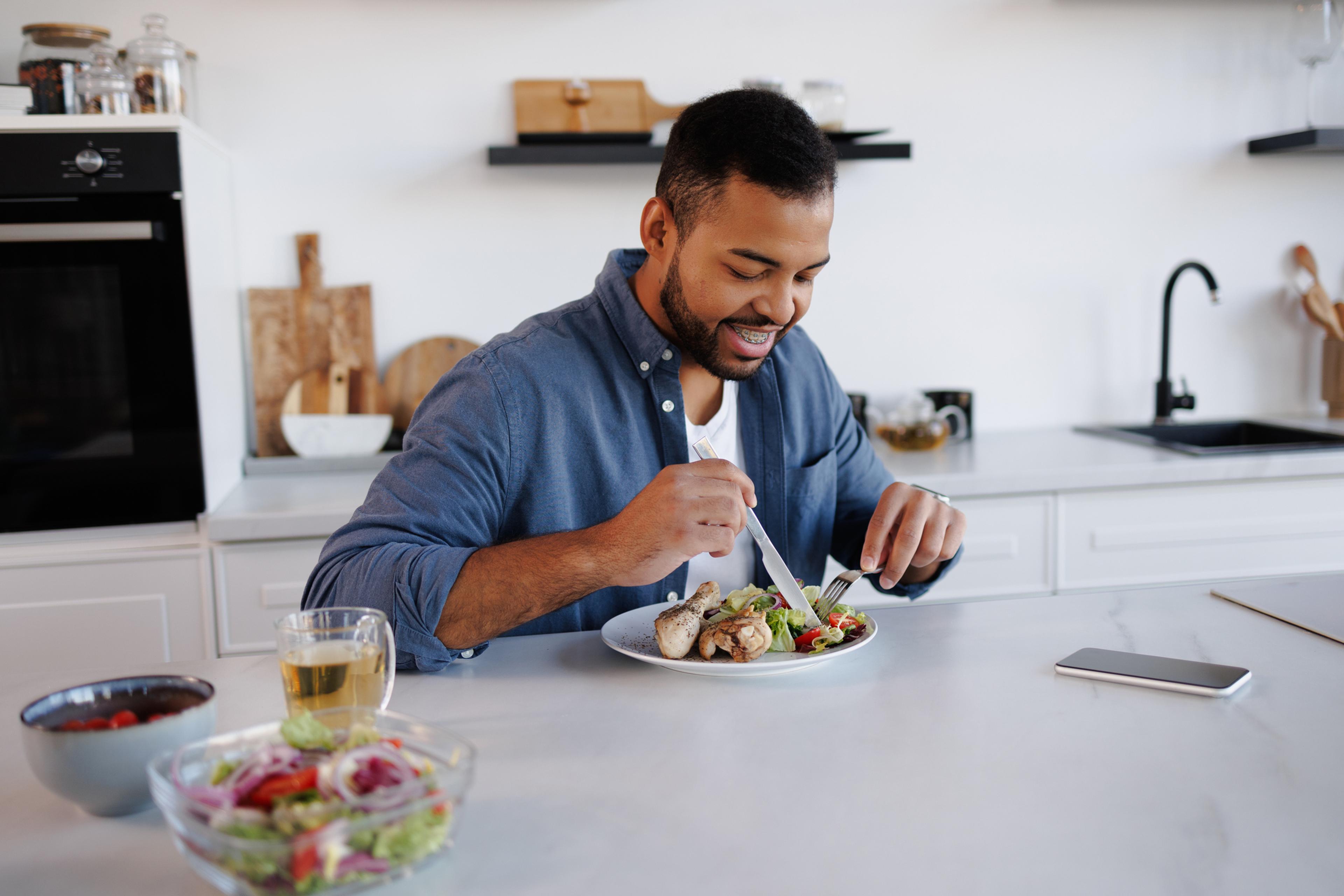 African American man smiling and eating fresh salad and chicken meat near tea and smartphone in his kitchen.