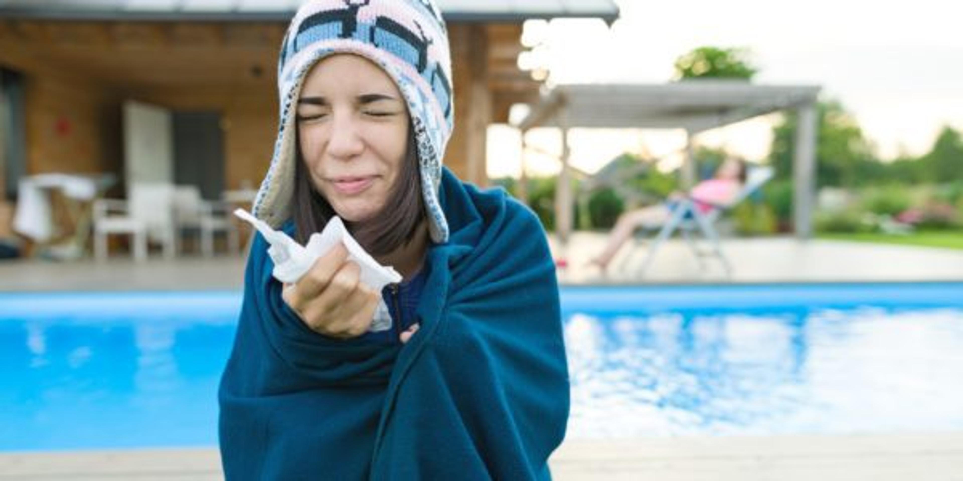 Flu, colds in the summer. Girl in a knitted hat with plaid with handkerchief sneezes, wipes her nose. Background nature, pool, girl in chaise longue