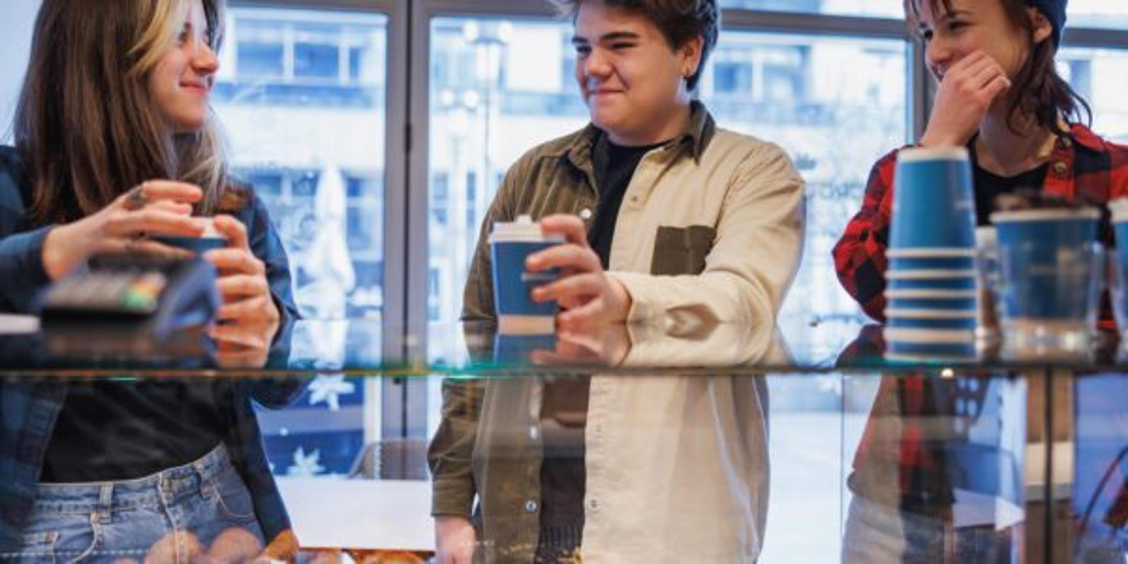 Cheerful teenagers standing by the baked pastry showcase counter, having coffee in paper cups